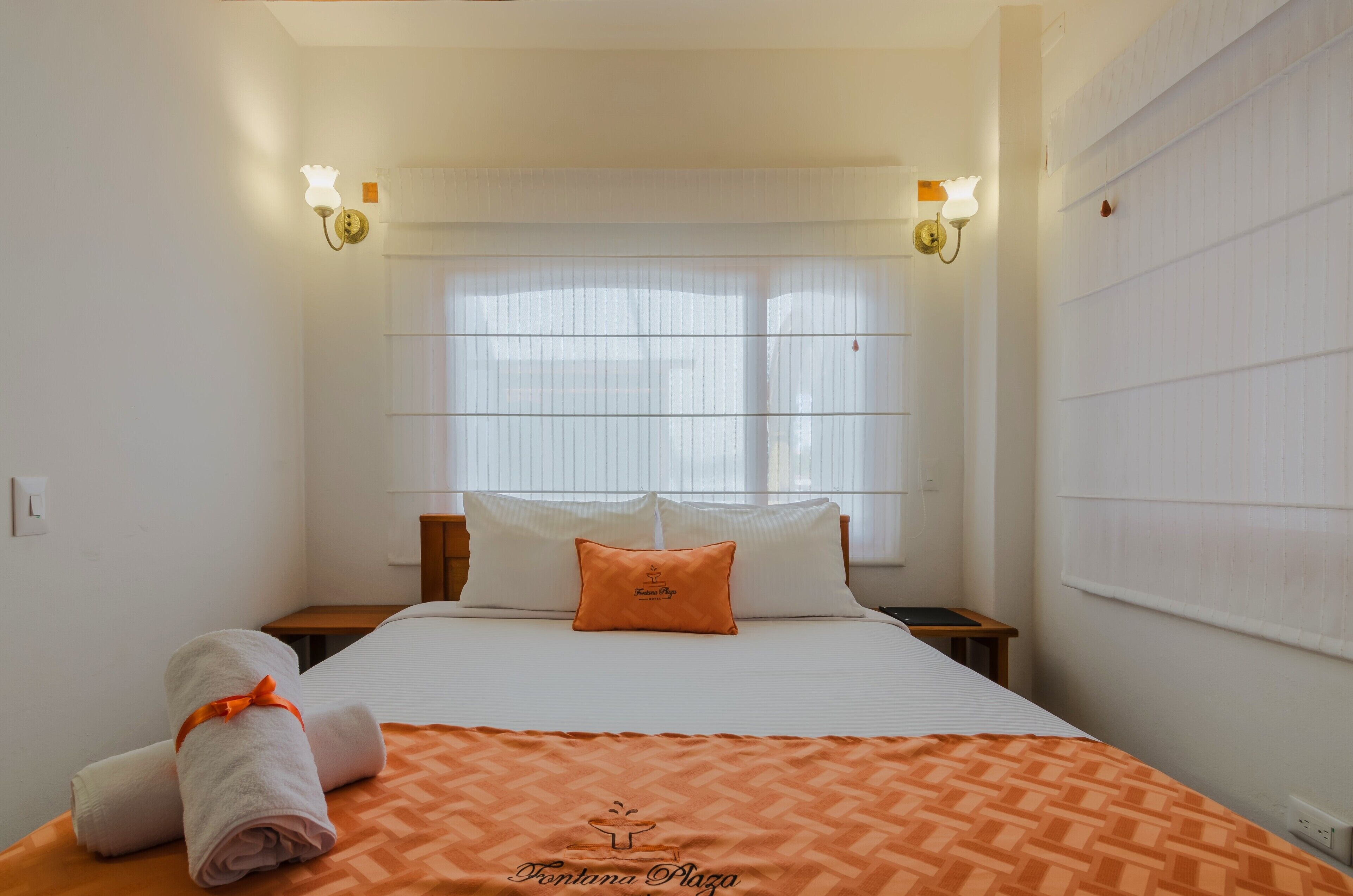 A hotel room with a bed, two bedside tables, a window with blinds, and wall-mounted light fixtures.