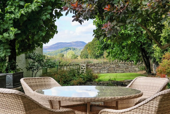 A garden patio with wicker furniture, a stone wall, and a view of a green landscape with hills.