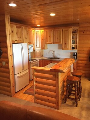 Une cuisine rustique aménagée dans une cabane en rondins, avec des meubles de rangement en bois, un réfrigérateur, un évier et un coin bar doté de tabourets.