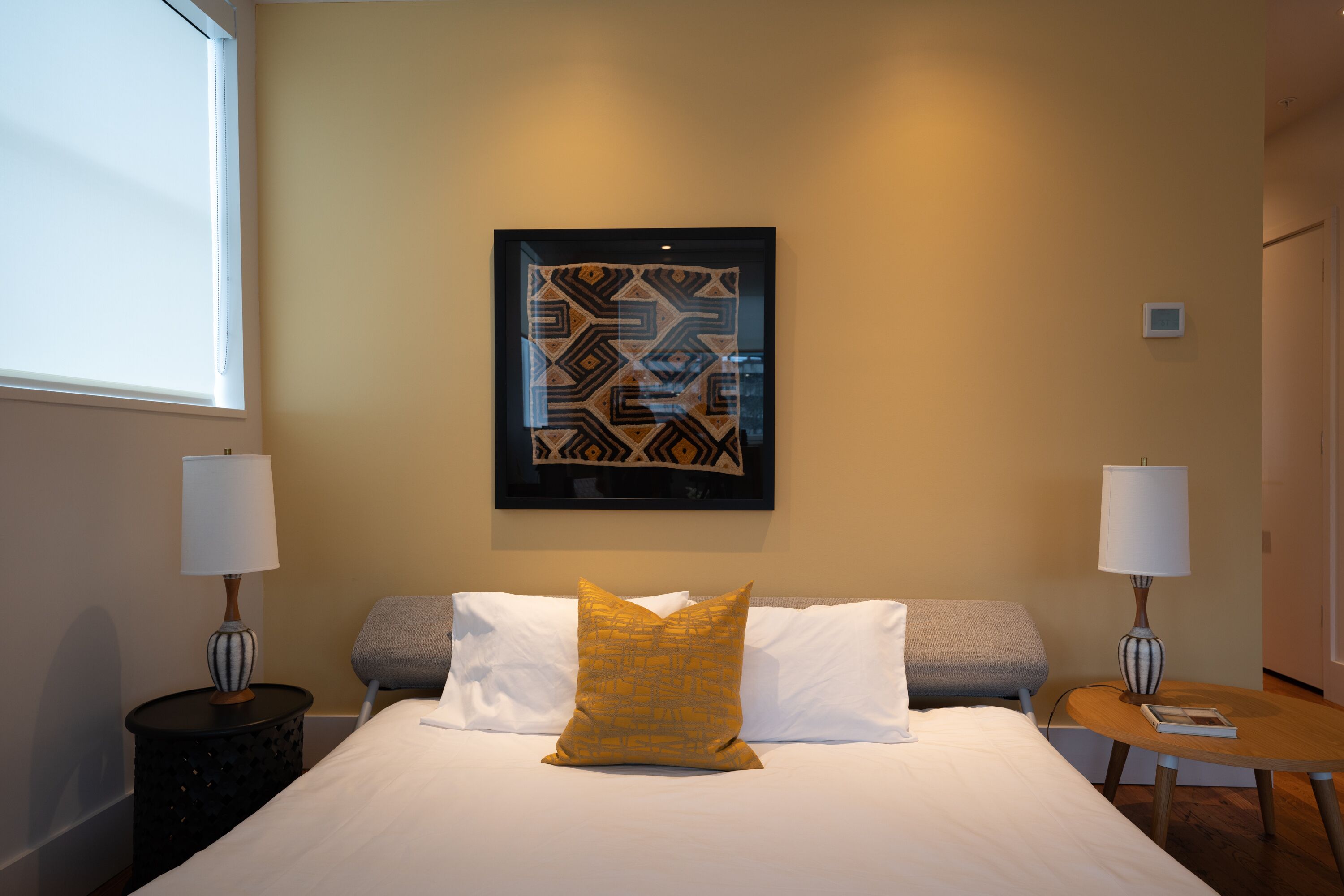 A hotel room with a bed, two bedside tables, a lamp on each table, a framed artwork above the bed, and a window on the left.
