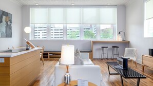 Una habitación de hotel moderna con suelo de madera, un sofá blanco, una mesa redonda con una lámpara, un banco y un escritorio con una computadora.