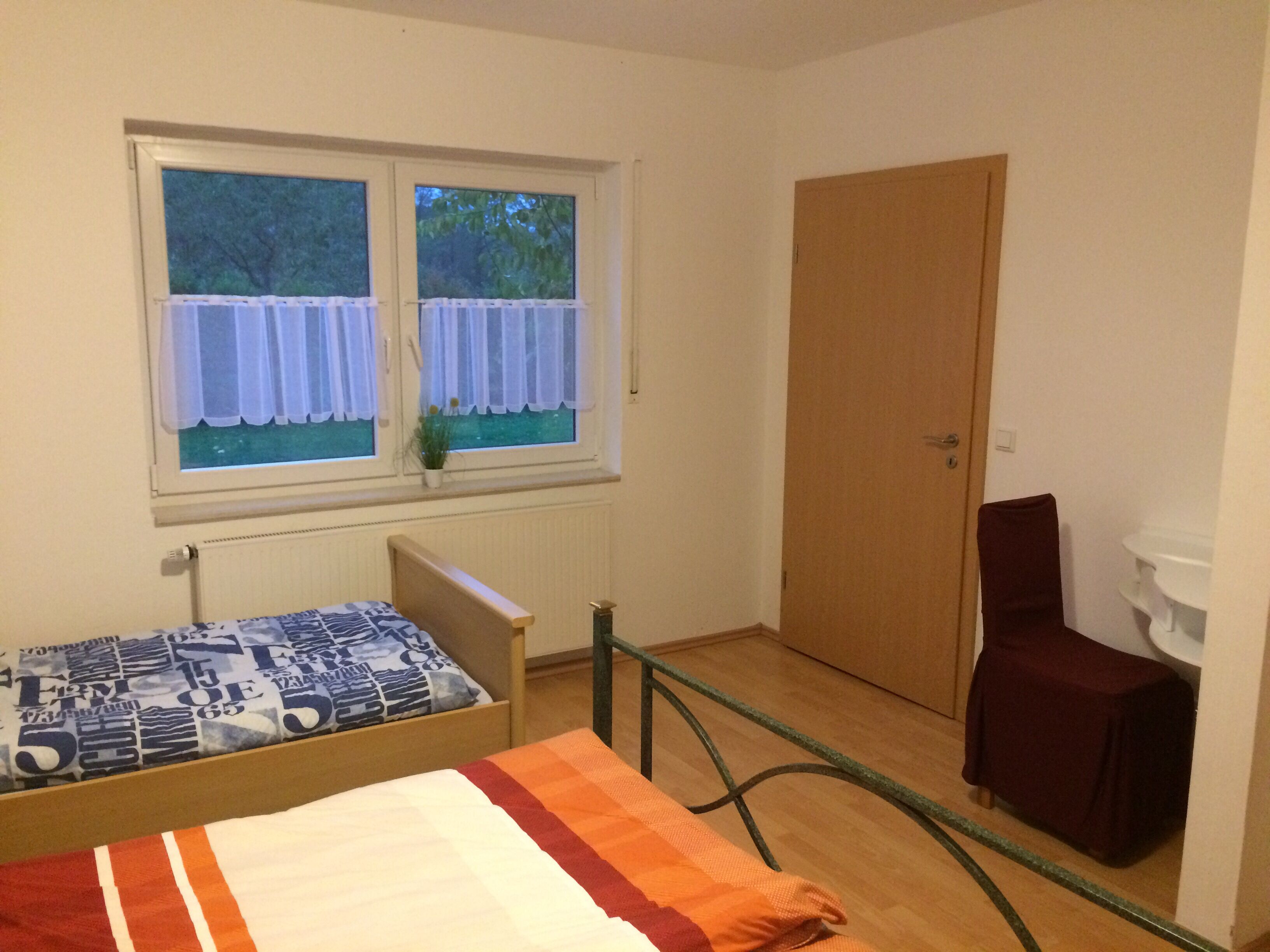 Una stanza piccola e modesta, con un letto, una finestra con tende, un termosifone e una porta.