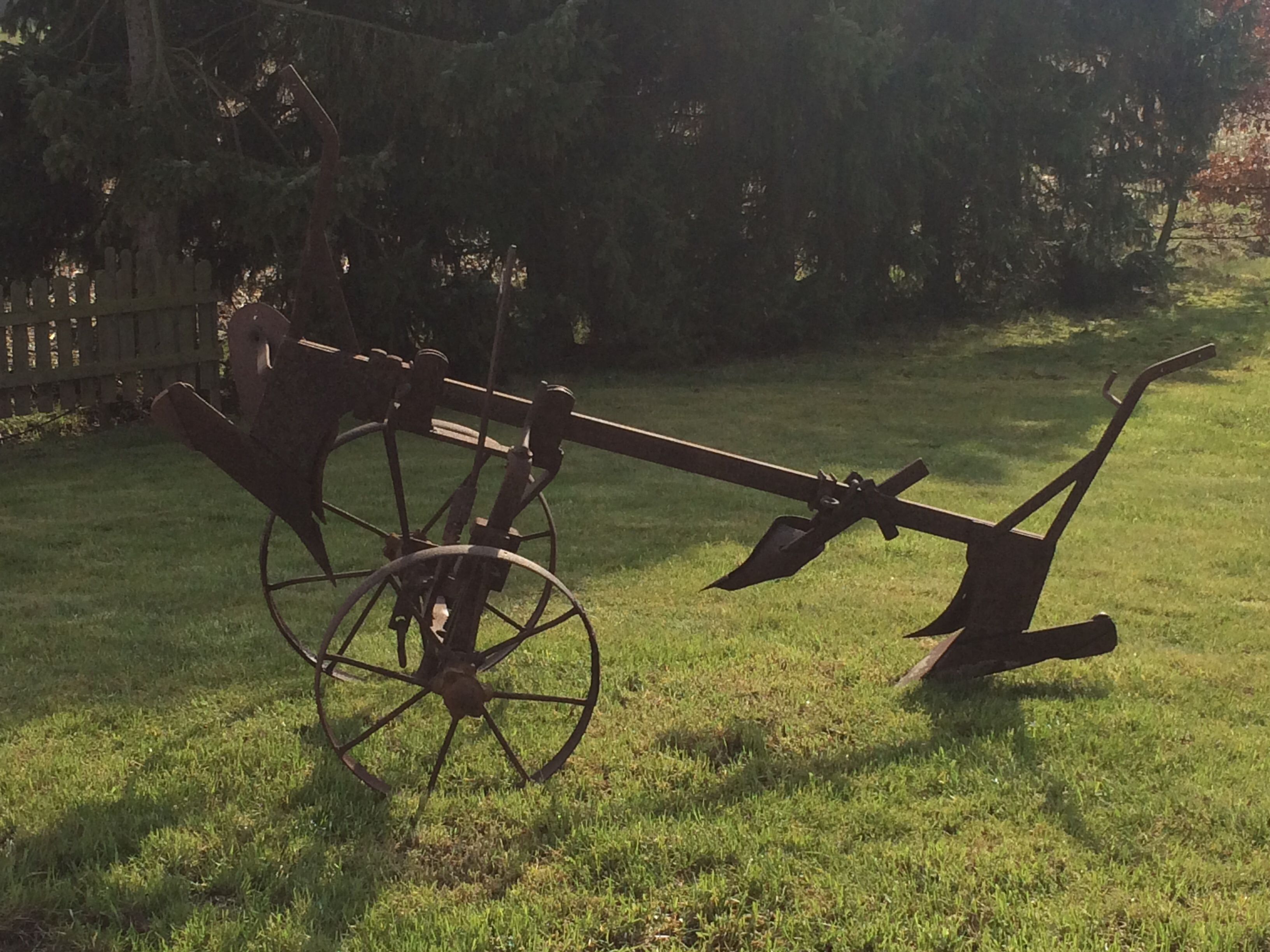 A vintage plow in a grassy field with a wooden fence in the background.