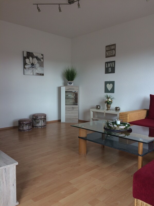 Ein modernes Wohnzimmer mit Glastisch, Holzboden, einem roten Sofa und Dekorationselementen wie einer Pflanze und Wandkunst.