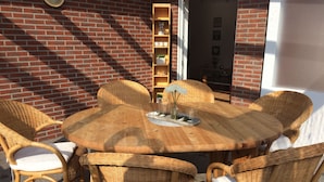 Une table ronde en bois, entourée de six chaises en osier, chacune agrémentée d’un coussin blanc. La scène se déroule à l’extérieur, avec un mur de briques et une étagère en bois en arrière-plan.