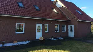 Une maison avec un toit recouvert de tuiles rouges, dotée de deux fenêtres et d’une porte, un petit jardin agrémenté d’un banc en bois, et un ciel dégagé.