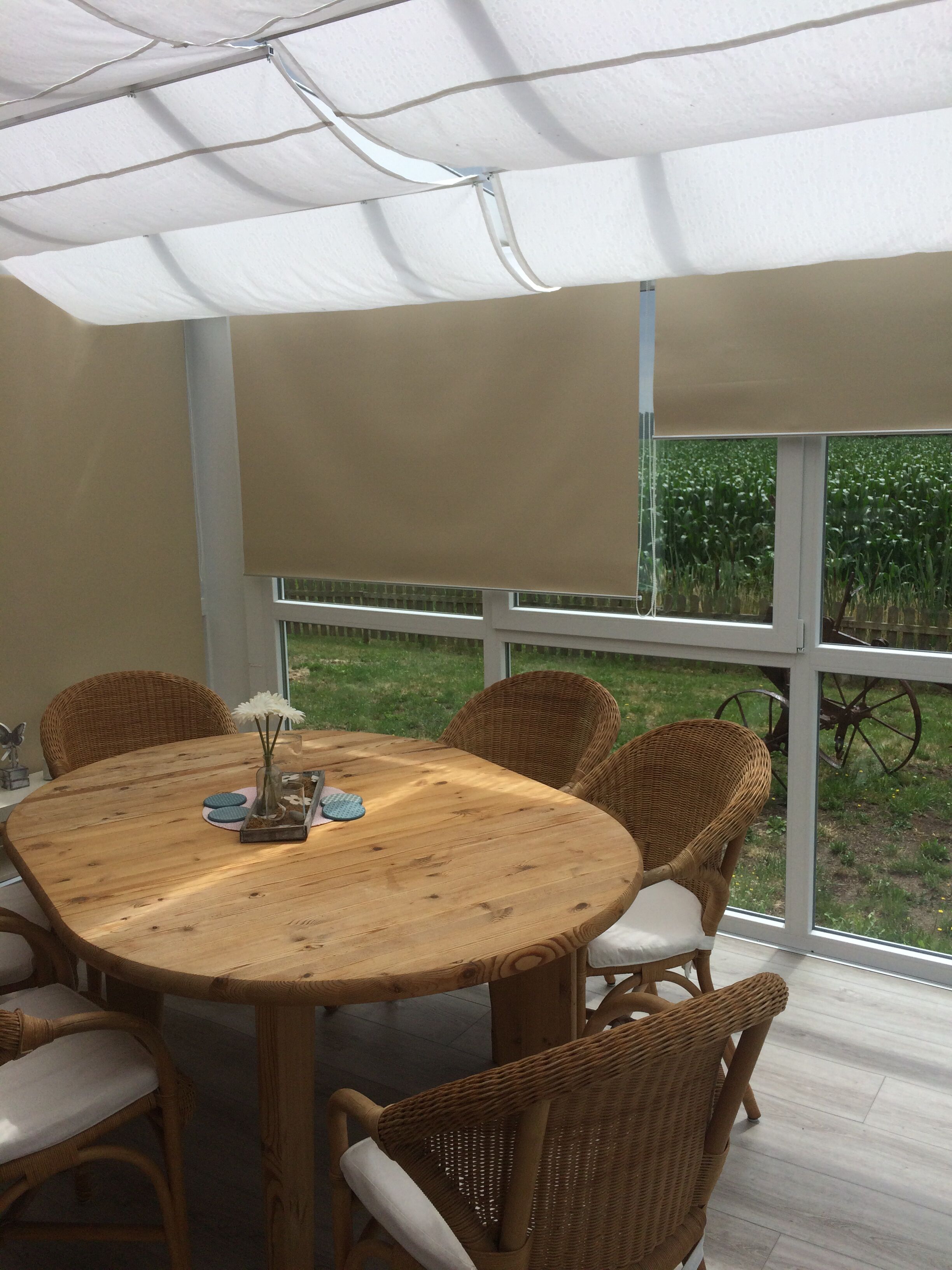 A covered outdoor dining area with a wooden table and wicker chairs, large windows, and a view of a green field.
