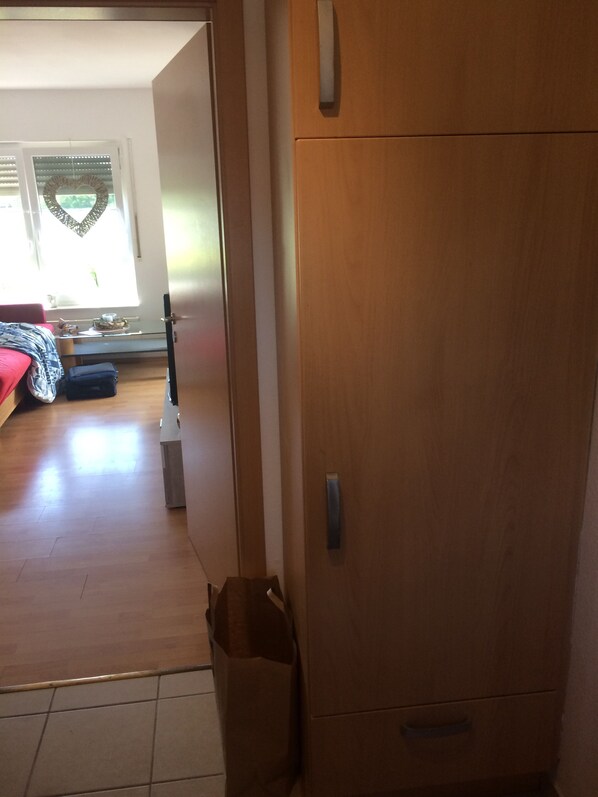 A wooden wardrobe with a handle, a tiled floor, and a visible door leading to a room with a bed and a window with blinds.
