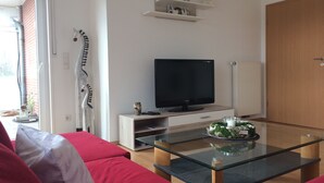 Un salon moderne avec un téléviseur à écran plat posé sur un meuble en bois, une table basse en verre et un canapé rouge. Une porte se trouve à droite et une fenêtre à gauche.