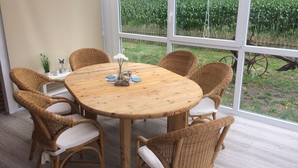 Une table ronde en bois, entourée de six chaises en osier, un vase rempli de fleurs et une vue sur un champ de maïs, visible à travers de grandes fenêtres.