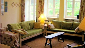 A cozy living room with two green sofas, a small table, and a lamp. There are large windows with curtains and a chandelier.