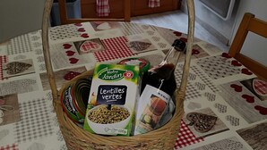 Una cesta de mimbre sobre una mesa con mantel a cuadros, que contiene un paquete de lentejas, una botella de salsa y un tazón pequeño de lentejas.