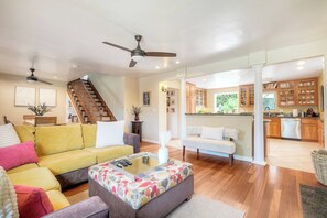 Amplia sala de estar con un sofá amarillo, un reposapiés gris y un sofá blanco. Una escalera conduce a una cocina con armarios de madera y un ventanal grande.