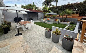 Une terrasse avec une table et des chaises, un barbecue, un coin salon avec des coussins et des plantes en pot.