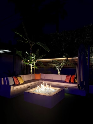 A nighttime outdoor seating area with a fire feature, cushioned seating, and surrounding greenery.