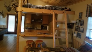 Ein rustikales Blockhaus-Interieur mit Schlafkoje, Küchenzeile, Holzwänden und einem aufgehängten Hirschkopf.