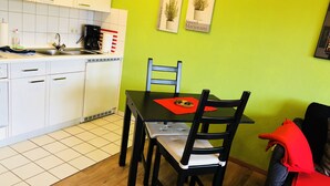 Una cocina compacta con armarios blancos, encimera negra y un lavamanos. Hay una mesa de comedor con dos sillas y un mantel individual rojo. Dos plantas en macetas decoran la pared.