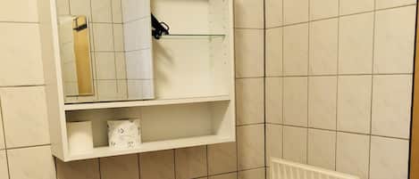 A bathroom with a toilet, a white radiator, a shelf with a mirror, and a roll of toilet paper.