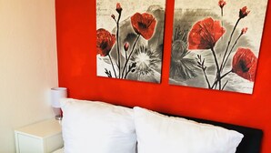Una cama con ropa de cama y almohadas blancas, una mesita de noche blanca con una lámpara, y dos cuadros florales rojos y blancos en la pared.
