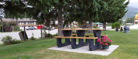 Une table de pique-nique avec des bancs, située dans un espace verdoyant avec des arbres, et un parking en arrière-plan.
