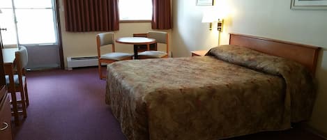 A hotel room with a bed, a desk, a chair, a small table, and a lamp.