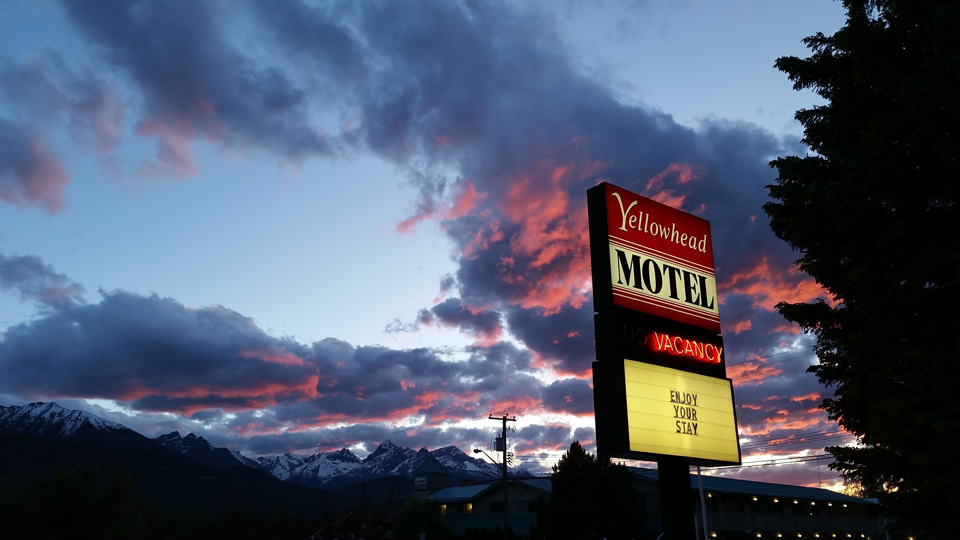 Ein Schild eines Motels mit der Aufschrift 'Keine Zimmer frei', vor einem dramatischen Sonnenuntergangshimmel und schneebedeckten Bergen.