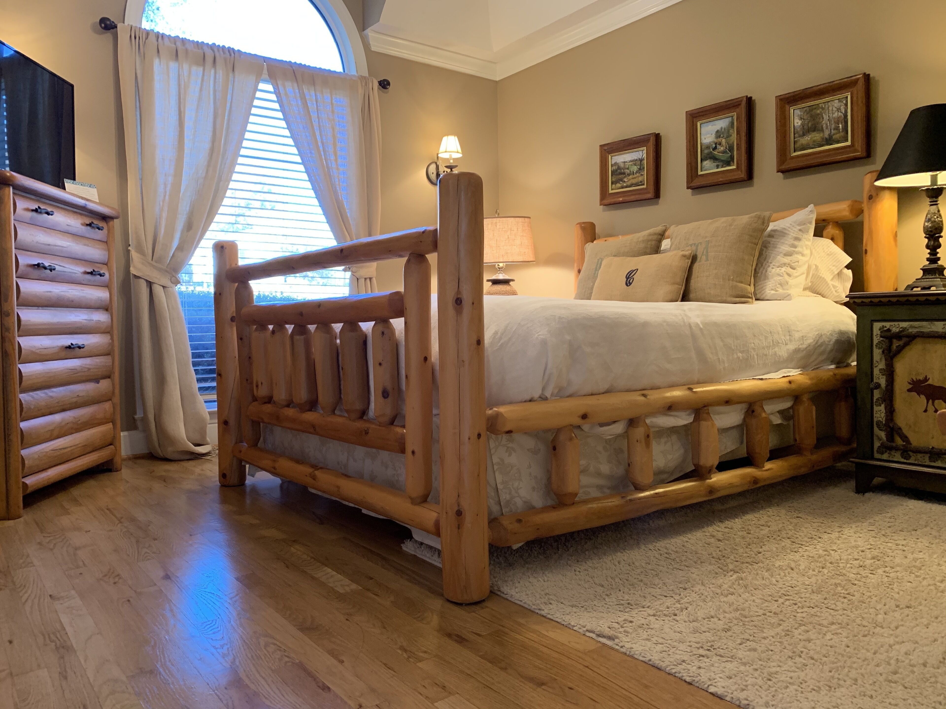 A bedroom with a wooden bed frame, a dresser, a lamp, and framed pictures on the wall.