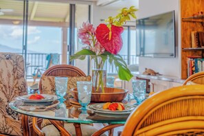 Un comedor con mesa de vidrio, sillas de mimbre, un florero con flores y vistas al exterior.