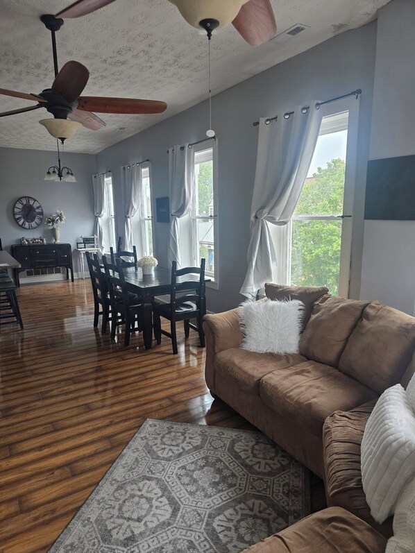 A living room with a brown sofa, wooden dining table, chairs, a ceiling fan, and large windows with curtains.