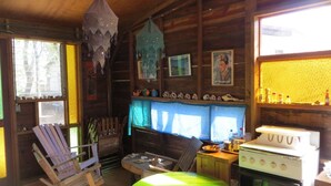 Interior rústico de una cabaña con paredes de madera, una mesa verde, un sillón mecedor, una estufa y una ventana con vistas a árboles.