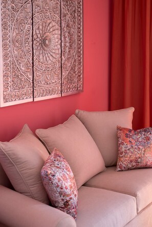 Um sofá bege com almofadas florais em frente a uma parede vermelha, com um painel decorativo acima.