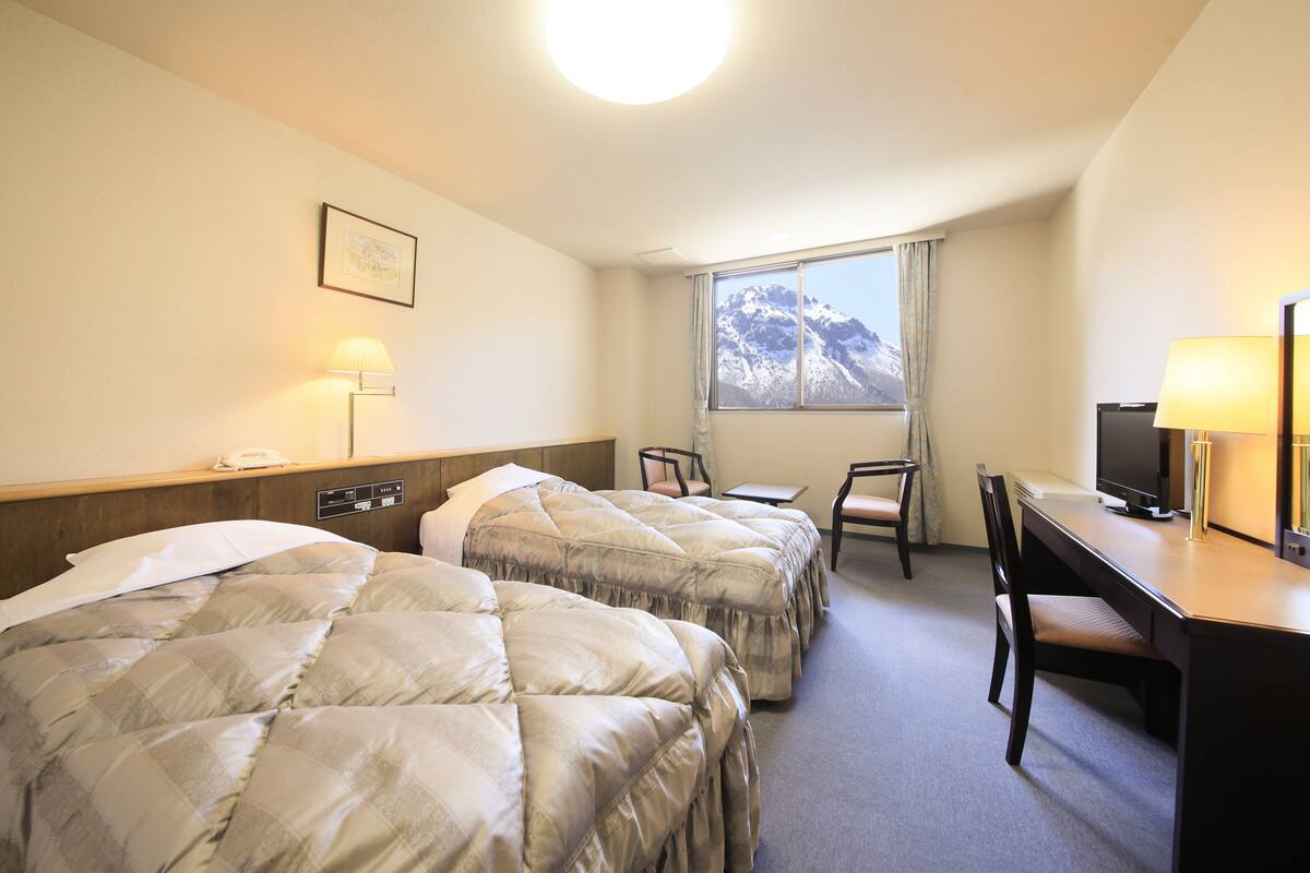 Ein Hotelzimmer mit zwei Betten, einem Schreibtisch, einem Sessel, einem Fernseher und einem Fenster mit Blick auf die Berge.