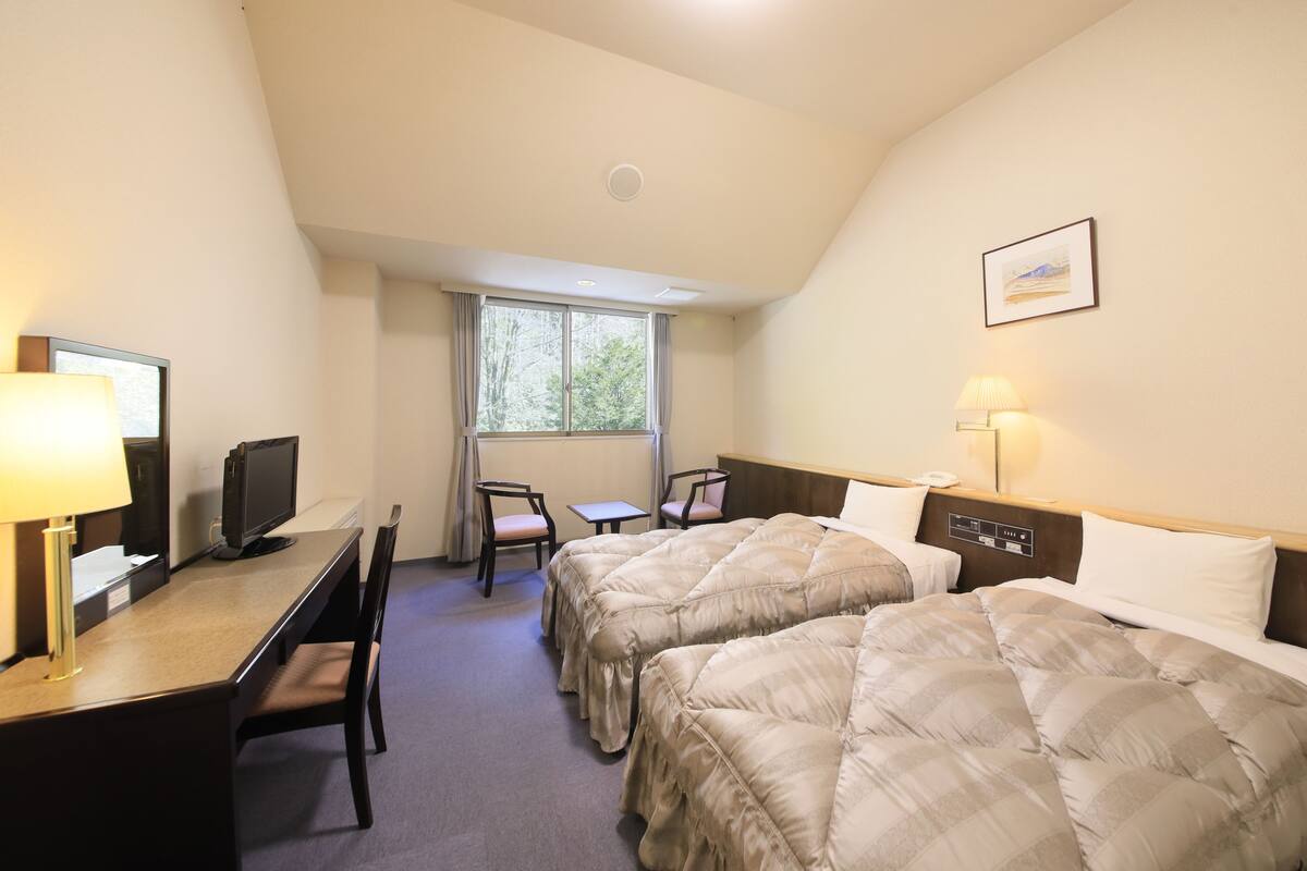 Ein Hotelzimmer mit zwei Betten, einem Schreibtisch mit Computer, einem Stuhl, einem Vorhangfenster und einer Wandlampe.