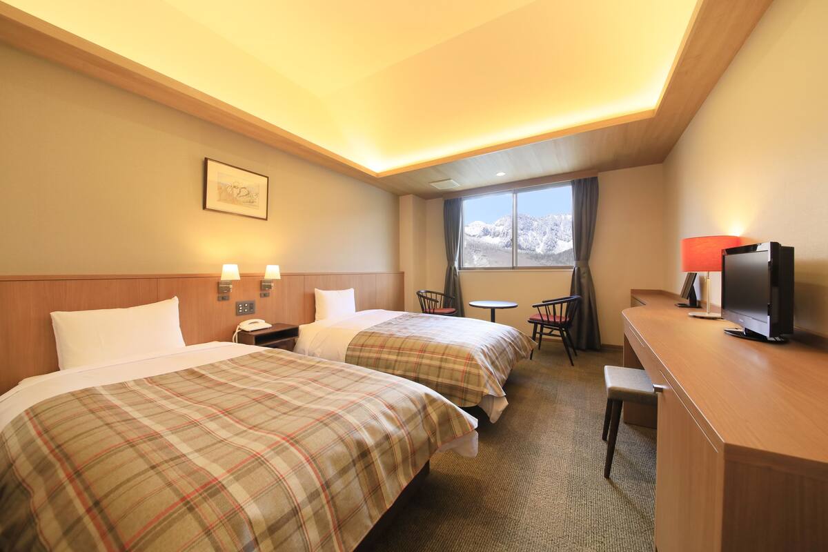 Ein Hotelzimmer mit zwei Betten, einem Schreibtisch, einem Sessel, einem Fernseher und einem Fenster mit Blick auf die Berge.