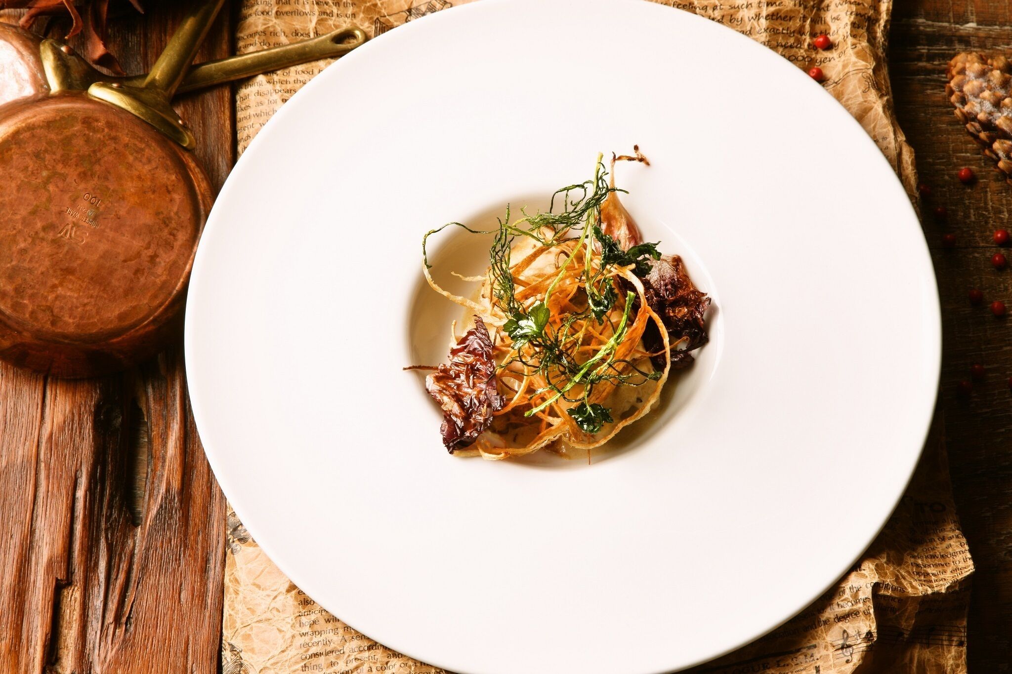 A white plate with a dish of food, a copper pot, and a wooden surface with newspaper.