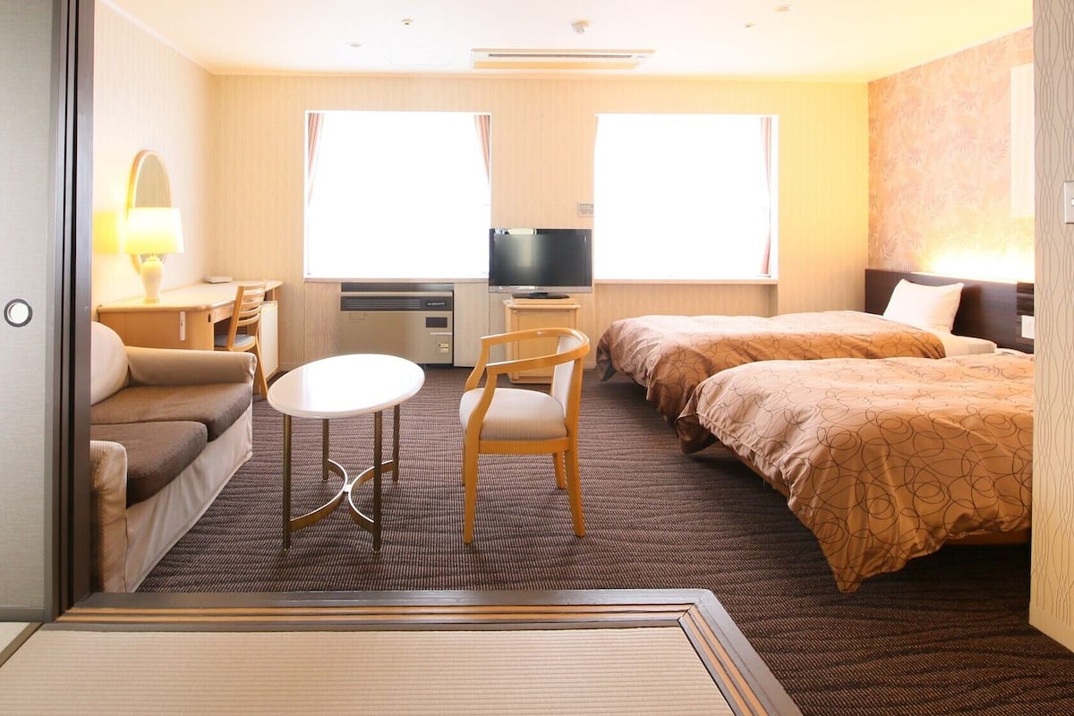 Una habitación de hotel con sofá, una mesita redonda, dos camas individuales, televisión y ventana.