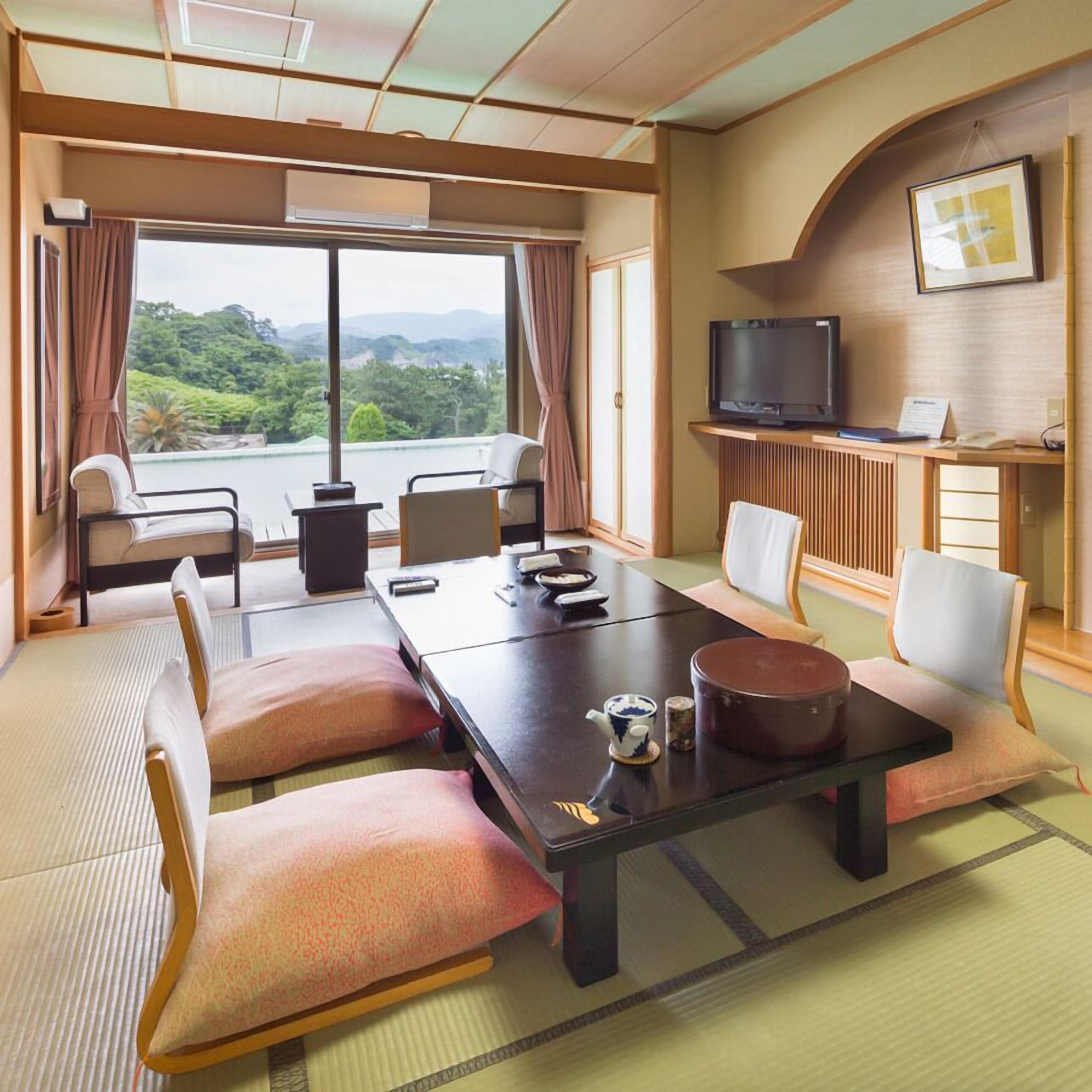 Camera d'albergo con un ampio tavolo da pranzo, sedie, televisione e vista su aree verdi.