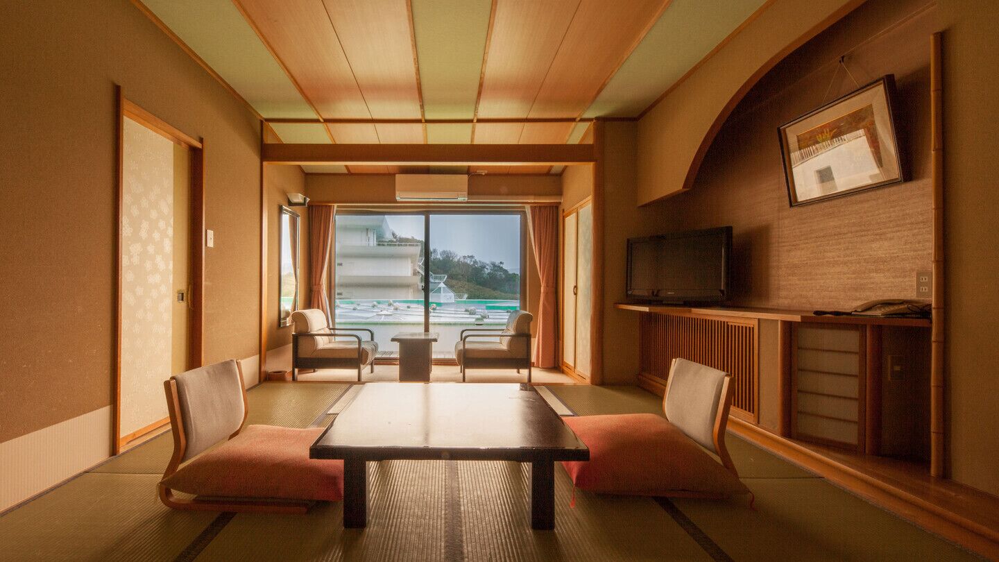 Una tradizionale stanza giapponese con pavimento in tatami, un tavolino, due poltrone imbottite, un televisore e vista su un edificio e un paesaggio attraverso un'ampia finestra.