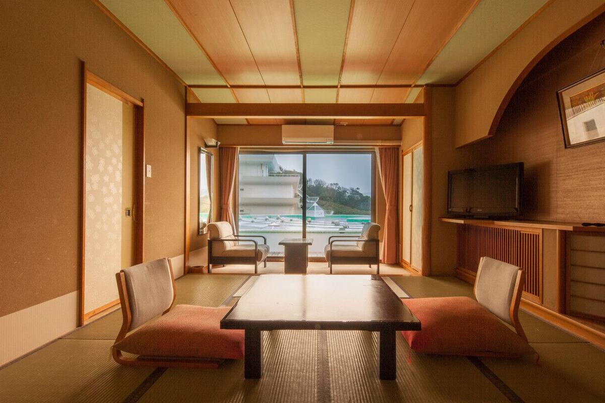 Une pièce de style japonais traditionnel, avec un sol recouvert de tatamis, une table basse, deux fauteuils rembourrés, un téléviseur et une vue sur un bâtiment et un paysage, le tout visible à travers une grande fenêtre.