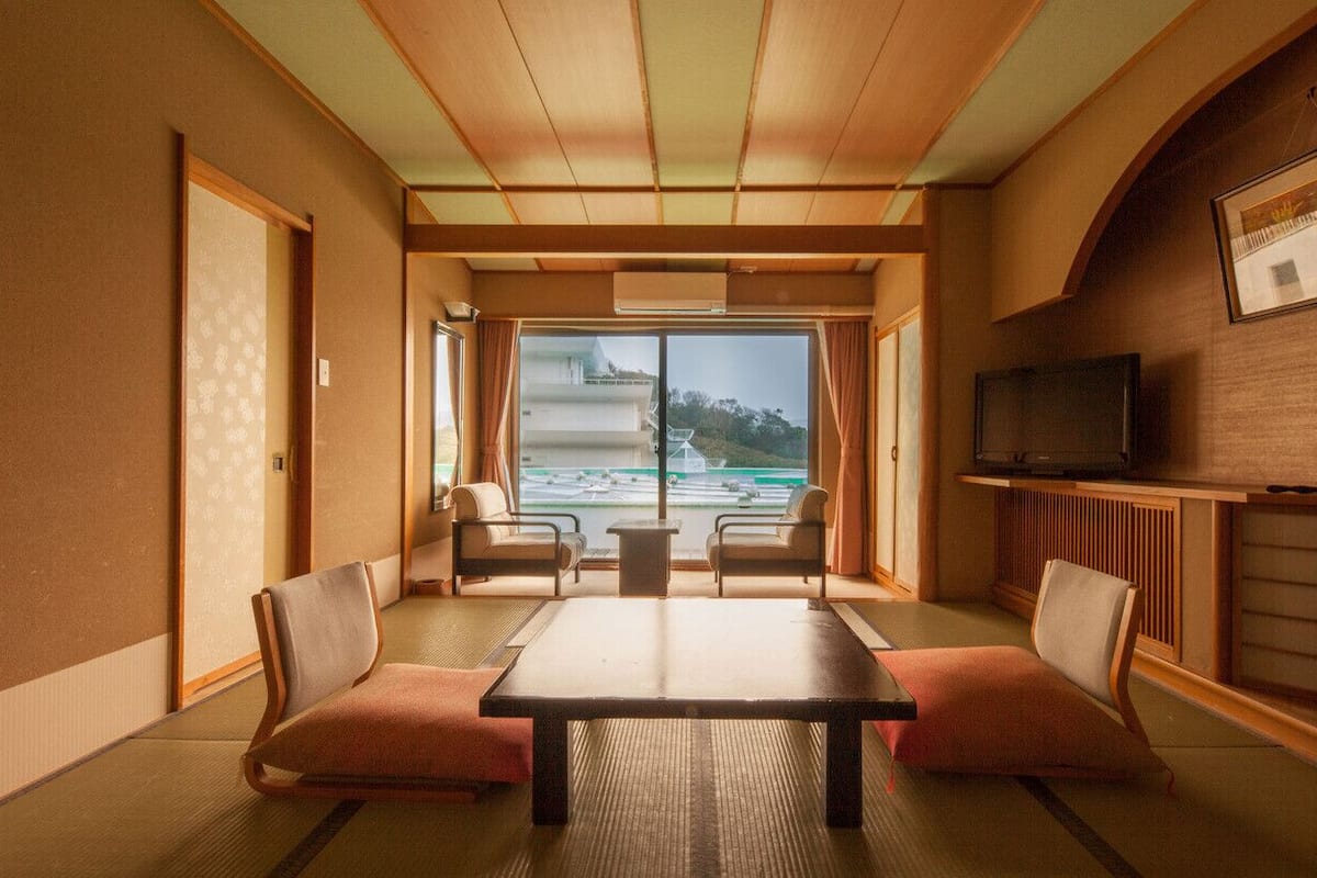 Una habitación japonesa tradicional con suelo de tatami, una mesa baja, dos sillas con cojines, un televisor y vistas a un edificio y paisaje a través de un amplio ventanal.