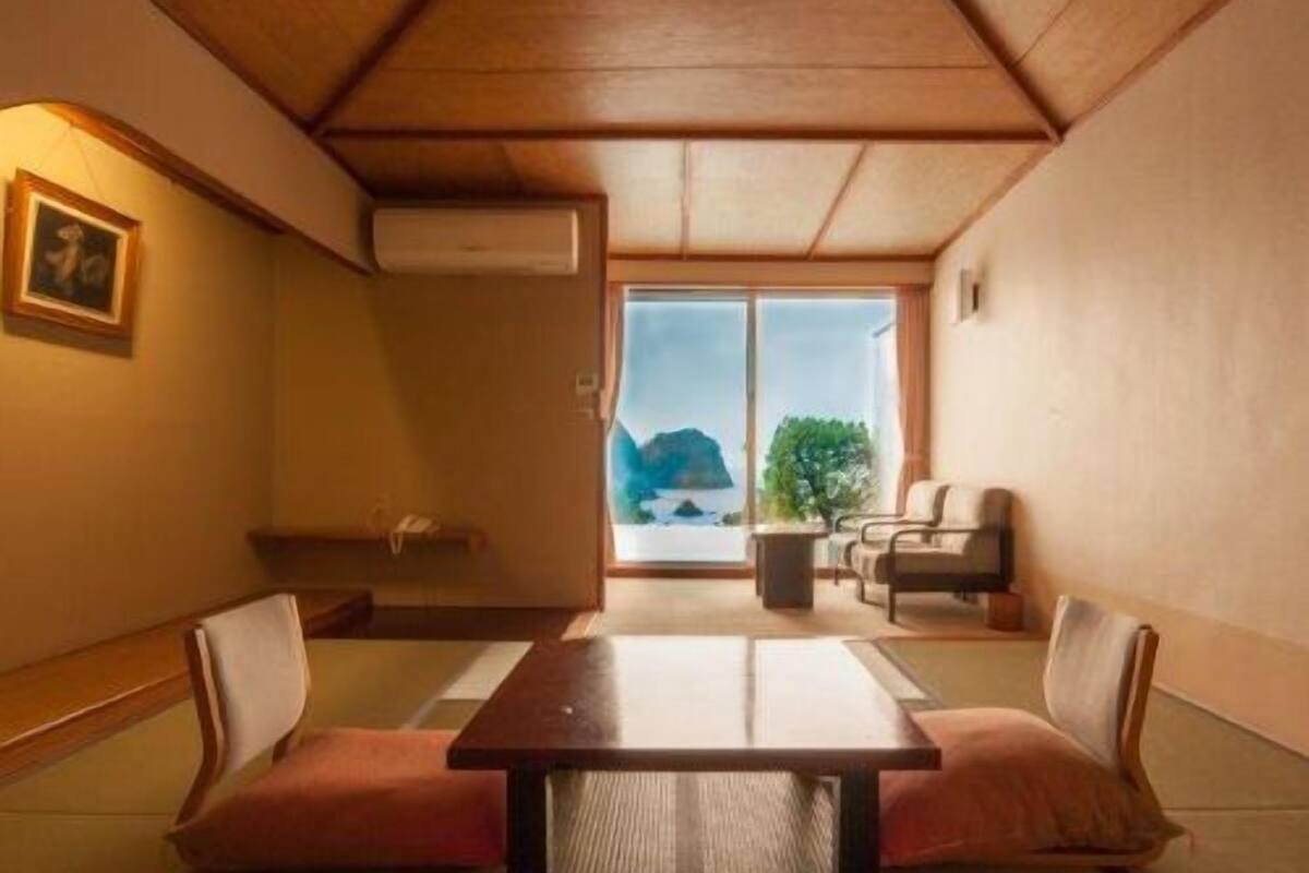 Une pièce traditionnelle avec un sol recouvert de tatamis, un plafond bas, une porte coulissante en verre donnant sur une vue sur la montagne, et une petite table avec des coussins.