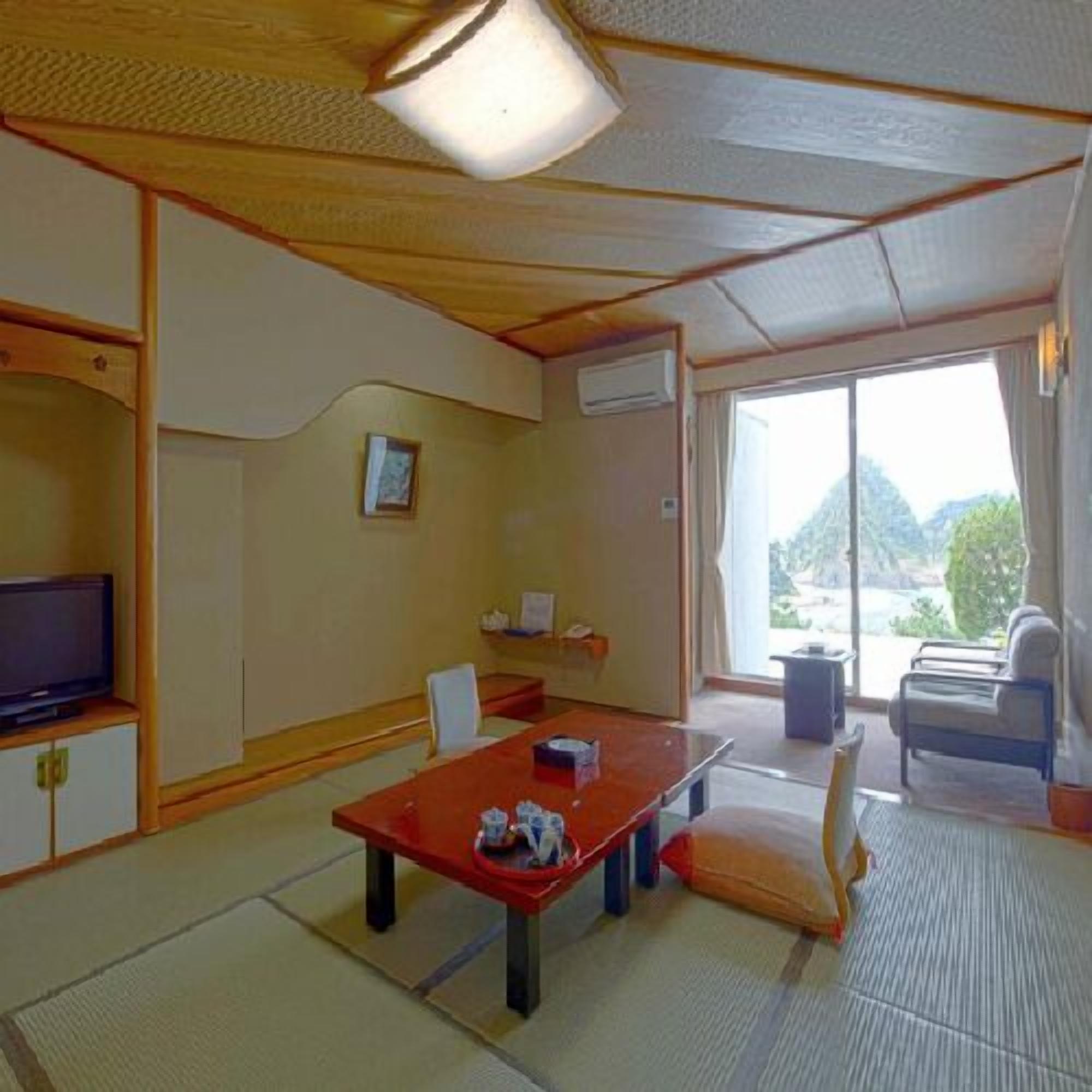 Una habitación tradicional japonesa con suelo de tatami, una mesa baja, televisión y vistas al exterior.
