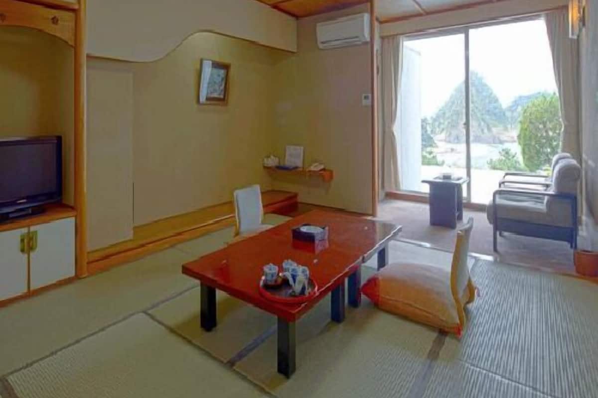 Une pièce de style japonais traditionnel, avec un sol recouvert de tatamis, une table basse, un téléviseur et une vue sur l’extérieur.