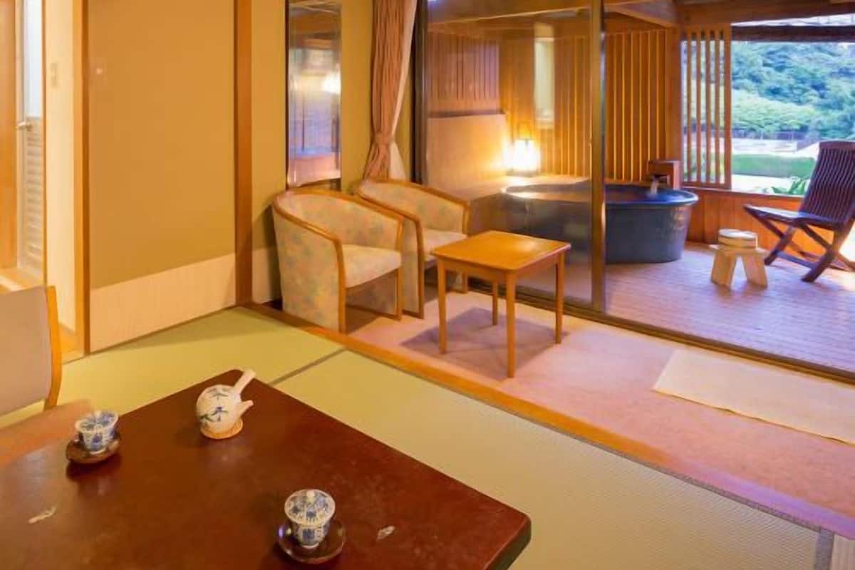 Una habitación tradicional japonesa con suelo de tatami, una mesa baja con juegos de té, una silla y un puertas correderas que dan a un espacio al aire libre.
