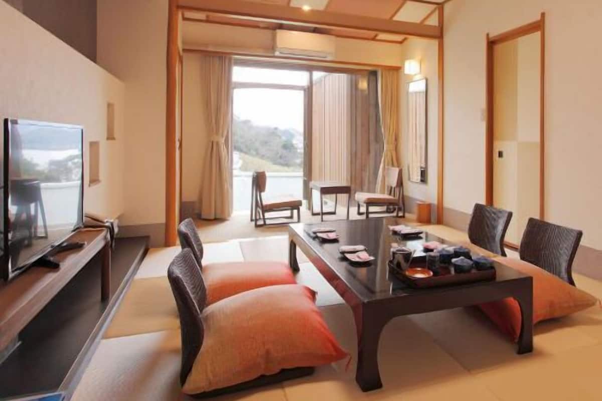 Une pièce de style japonais traditionnel, avec un sol recouvert de tatamis, une petite table basse avec des coussins, un téléviseur et une vue sur l’extérieur.