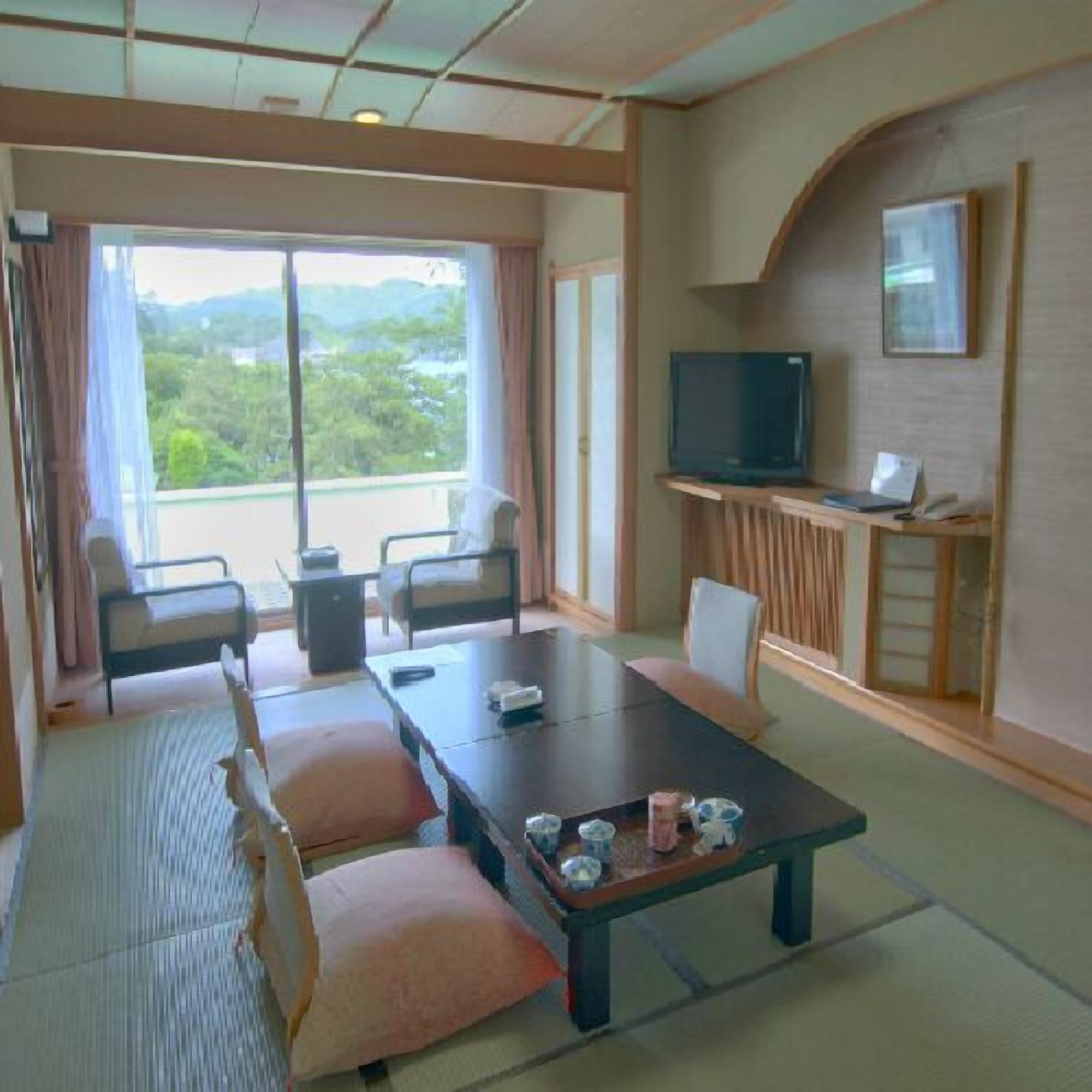Camera d'albergo con un'ampia porta-finestra, una televisione su un mobile, un tavolo da pranzo con sedie e vista su spazi verdi.