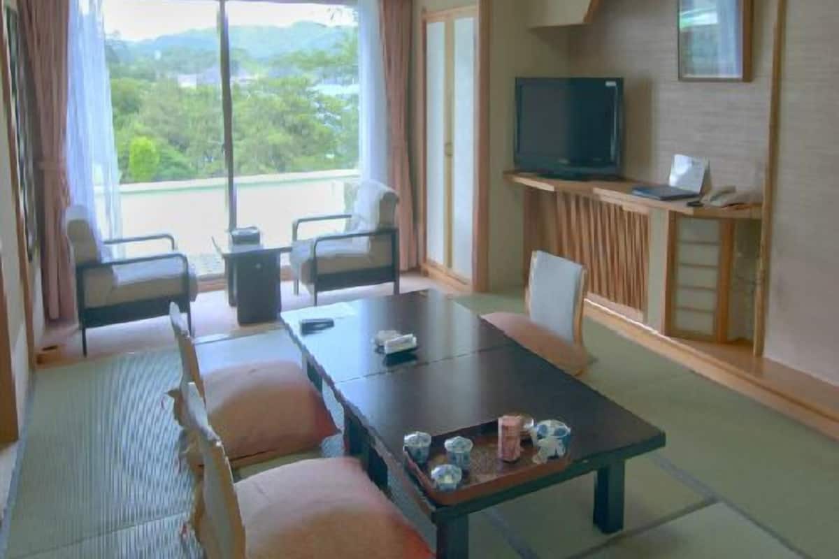 Une chambre d’hôtel dotée d’une grande porte-fenêtre coulissante, d’un téléviseur posé sur un meuble, d’une table à manger avec des chaises, et offrant une vue sur la verdure environnante.