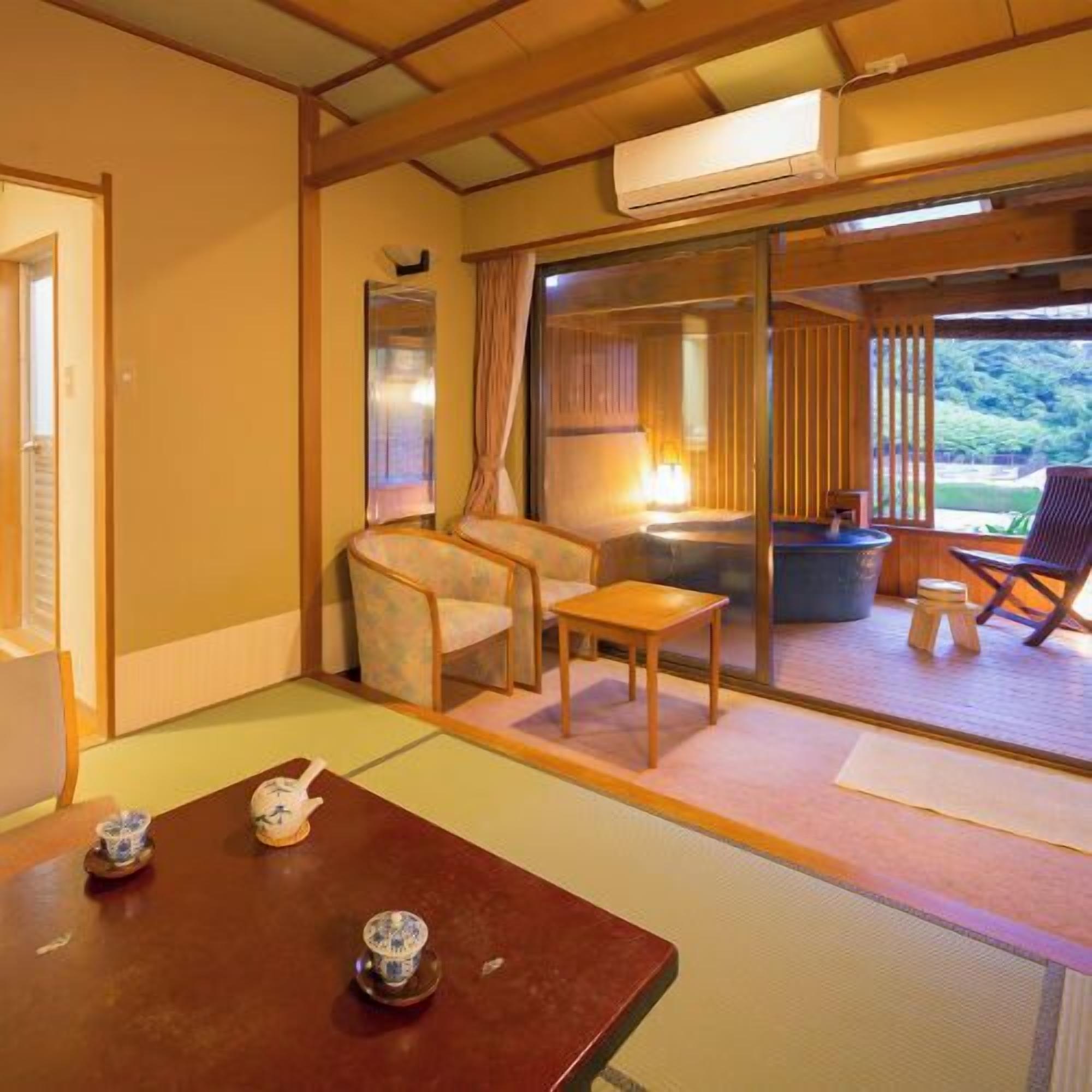 Una habitación japonesa tradicional con suelo de tatami, una mesa baja con juego de té, una silla y vistas a un área exterior con mesa y sillas a través de una puerta corredera.