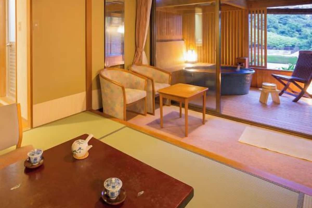 Une pièce de style japonais traditionnel, avec un sol recouvert de tatamis, une petite table avec un service à thé, une chaise, et une vue sur un espace extérieur aménagé avec une table et des chaises, accessible par une porte coulissante.