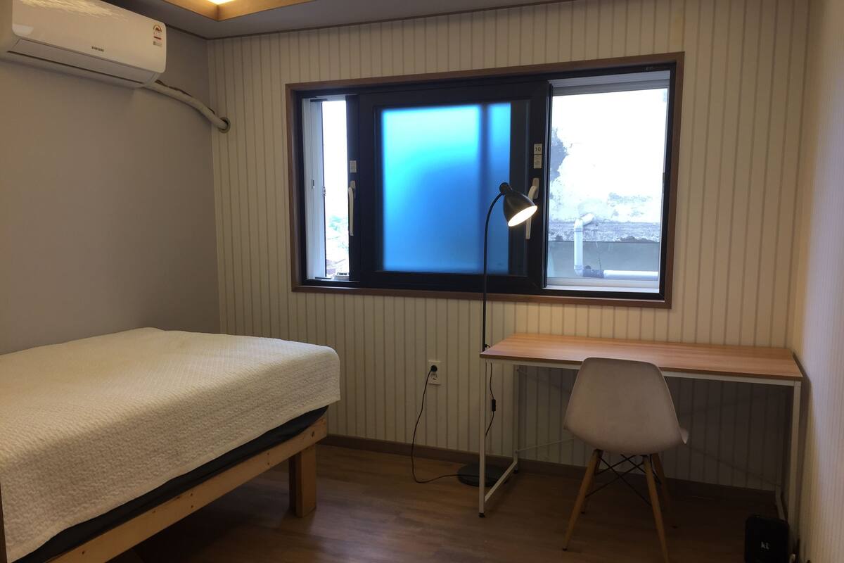 Une chambre avec un lit simple, un bureau, une chaise, une fenêtre avec des stores et un appareil de climatisation.
